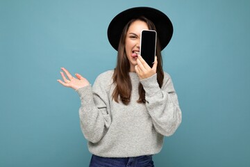 Sexy Photo of Beautiful positive woman wearing black hat and grey sweater holding mobilephone showing smartphone isolated on background looking at camera and showing tongue