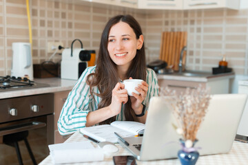 Happy young woman holding cup of coffee and sitting in front of laptop, working from home.