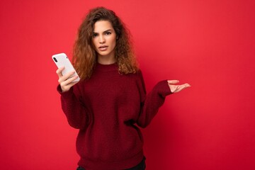 Photo of pretty asking upset young woman with curly hair wearing dark red sweater isolated on red...