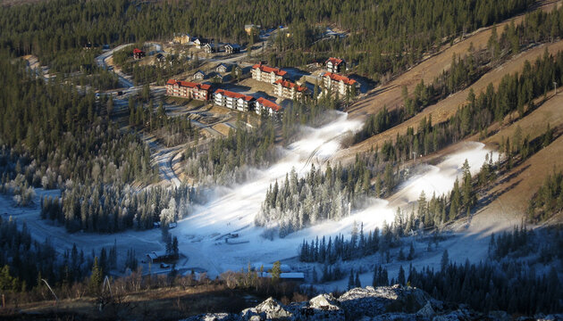 Beschneiung (technische Schneeerzeugung) in einem Skigebiet mit Skiliften und Skipisten. Im Hintergrund eine Hotelanlage. CO2 Fu&szlig;abdruck im Kontext von Klimawandel und globaler Erderw&auml;rmung.