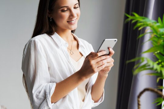 Portrait of business woman, holds smartphone in her hands. Beautiful girl uses mobile phone for work, calls clients, sends text messages. Millennial girl in white shirt stands against white wall