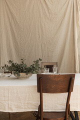 Modern interior dinning room design with bohemian styled decorations. Dinning table, eucalyptus branches, wineglasses, plates, cutlery, chair on linen tablecloth. Boho style concept.