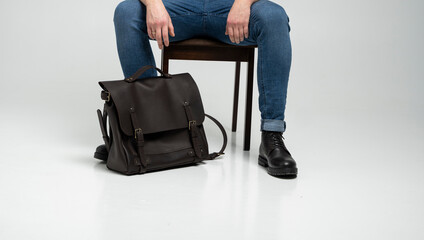 Man in a blue jeans and black boots sits on a chair with a brown men's shoulder leather bag for a documents and laptop on a white floor. Mens leather satchel, messenger bags, handmade briefcase.