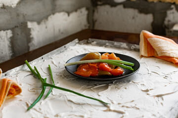 Pickled sweet bell pepper, cut into slices, multicolored vegetables in oil on a black matte plate and a stone white background. The dish is decorated with green leeks
