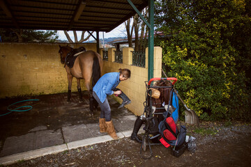Woman cleaning her riding boots with her brown horse in the background