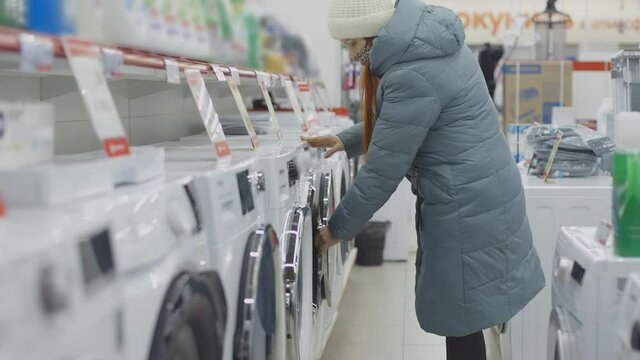 a woman in a medical protective mask chooses a washing machine in a home appliance store, observing social distance and precautions in a hypermarket during the epidemic, soft focus