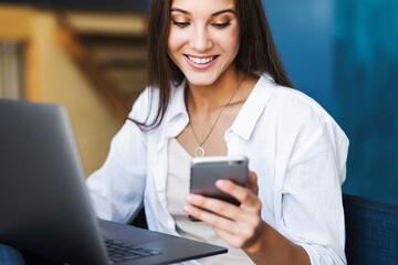 Business woman sits at table in front of laptop, writes a message using smartphone, conducts...