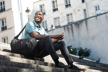 Full body young black man sitting on steps in city with cellphone and bag