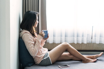Young happy Asian woman sitting on sofa and drinking hot coffee in the morning while looking...