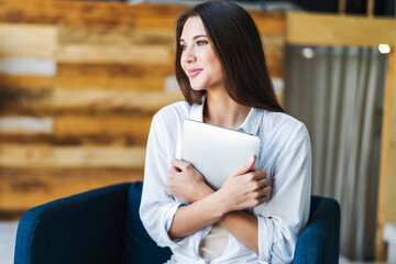 Smiling beautiful woman in white shirt sits on an armchair and squeezes smart tablet tightly. Millennial woman in good mood rejoices at work done.