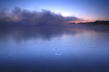 朝靄の漂う夜明けの湖。結氷した湖面に映る空の色。屈斜路湖、北海道、日本。