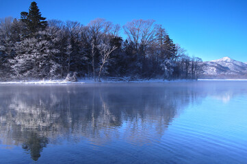 冬の青空の下の湖の風景。湖面に映る湖畔の森。阿寒摩周国立公園の屈斜路湖、北海道、日本。