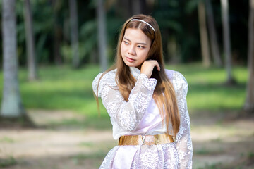 Asian woman in a Thai dress is standing in the garden with green tree background. Beautiful Thai girl looking something.