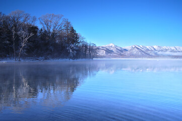 冬の青空の下の湖の風景。湖面に映る湖畔の森。阿寒摩周国立公園の屈斜路湖、北海道、日本。