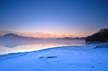 Fototapeta na wymiar 朝靄の漂う冬の朝の湖、雪に覆われた湖岸。夜明けの空のグラデーションと湖岸の森のシルエット。阿寒摩周国立公園の屈斜路湖。北海道、日本。