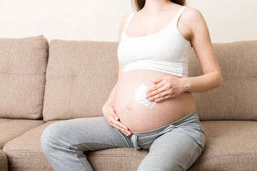 a pregnant girl sits at home on the bed and smears an anti-stretch mark cream on her stomach. Pregnancy, motherhood, preparation and expectation concept