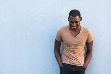happy young African American guy leaning against gray wall with hands in pocket and looking down