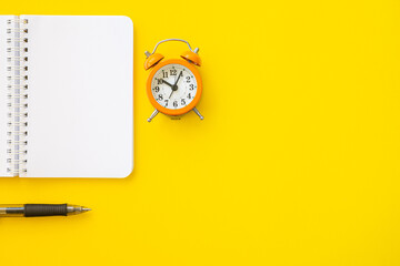 Top view of a notebook, a black pen and a small orange retro-style alarm clock. Isolated on yellow background with lot of copy space on the right.