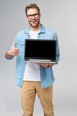 Concentrated young bearded man wearing glasses dressed in jeans shirt holding laptop isolated over grey studio background