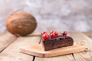A piece of gluten-free cake in chocolate, decorated with cranberries, on a blurry background. In the background is a coconut.