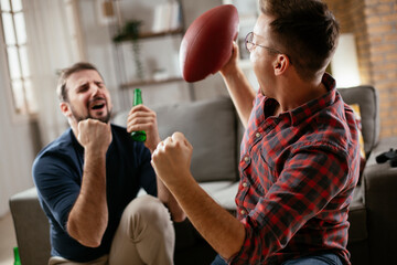 Two young friends enjoying at home. Men drinking beer and watching sports game on tv