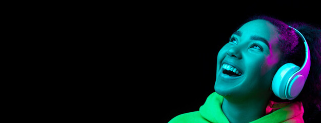 Laughting, flyer. African-american woman isolated on dark background in multicolored neon. Listening to music with headphones. Concept of human emotions, facial expression, sales, ad, fashion.