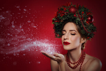 Beautiful young woman with Christmas wreath blowing kiss on red background