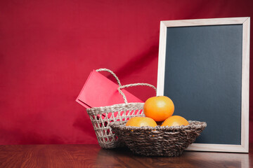 A small blackboard, four tangerines, and red envelopes (Ang Pao) in a basket placed on a wooden table with a red cloth background. Space for text. Happy Chinese New Year concept