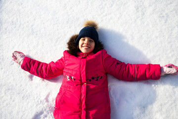Fototapeta na wymiar A cute little girl is lying in the snow and smiling. A child playing in the snow.