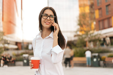Fototapeta premium Corporate Communication. Caucasian Businesswoman in eyewear Talking On Mobile Phone with coffee to go on business center background