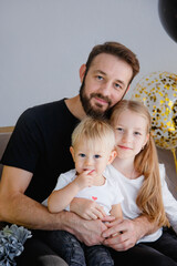 Portrait of a happy father with adorable children sitting on the sofa posing for a family photo at a holiday home. Cuddles with cute little son and daughter while looking at the camera