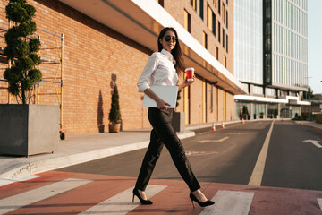 Businesswoman having a break from work and relaxing while crossing the street on zebra holding coffee to go with business center in background.