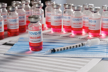 Vaccine vial, syringe and many glass bottles on background for vaccination against COVID-19 SARS-CoV-2 coronavirus pandemic lying on surgical mask and immunization document. 