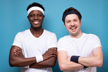 Smiling cheerful young two friends, european and african men standing and looking camera confident. Studio shot on blue wall.