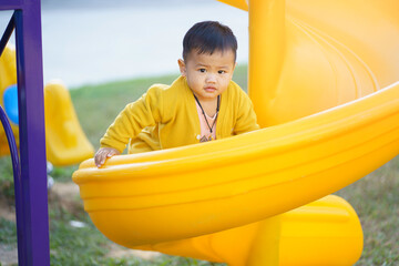A 2-year-old Asian boy wearing a yellow shirt and blue pants is happily playing with a slide at the playground, Happy children, The child is smiling and happy