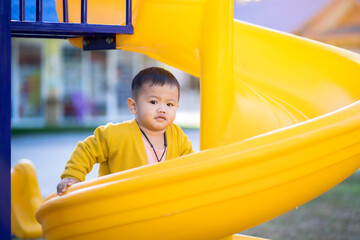 A 2-year-old Asian boy wearing a yellow shirt and blue pants is happily playing with a slide at the playground, Happy children, The child is smiling and happy