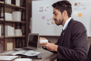 Fototapeta na wymiar Young smart businessman drinking coffee in working room. Handsome arabic businessman sitting and holding cup of coffee. Business and working concept.