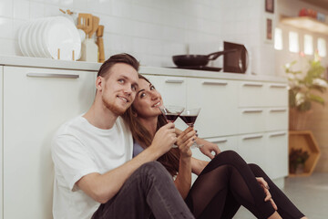 Happy couple love drinking wine together while cooking in the kitchen. Young loving man and woman sitting and drinking wine together in kitchen room.