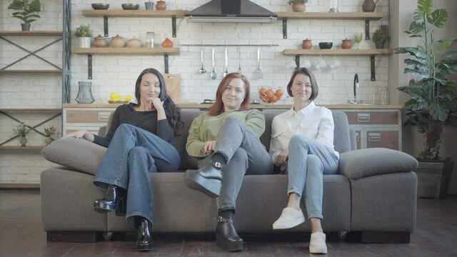 Wide shot portrait of three confident young Caucasian women sitting on couch crossing legs. Beautiful female friends enjoying leisure indoors. Confidence and femininity concept.