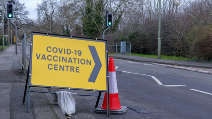 Temporary yellow directional sign showing way to a newly opened Covid-19 Vaccination Centre. Placed on pavement and road. With sandbag and cone. Open road. Landscape image, with space for text. UK. - 406726054