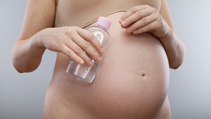 Pregnant woman holds body oil for stretch marks. Pregnant belly isolated on a grey background