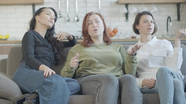 Relaxed Caucasian women sitting on couch watching TV and talking. Three carefree friends discussing movie indoors. relaxation and leisure concept.