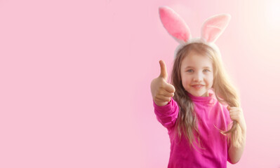 Obraz na płótnie Canvas Happy Easter. A cute little girl in a pink sweater with bunny ears smiles cutely and shows a thumb up on a pink background. Banner. Class sign. Horizontal format. Copyspace