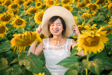 A lovely Asian woman in a white decent dress wearing a white hat surrounding by beautiful sunflower and enjoying her holiday in the park which is full of plants.