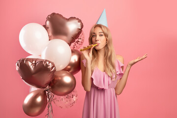 Obraz na płótnie Canvas Blonde woman with air balloons and birthday cap blowing party horn, celebrating her special day on pink background
