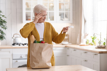 Shocked elderly woman checking grocery bill in kitchen - obrazy, fototapety, plakaty