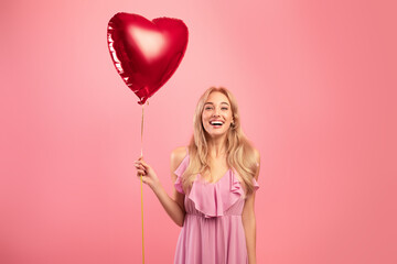 Portrait of joyful blonde woman in dress holding red heart shaped balloon for Valentine's Day on...