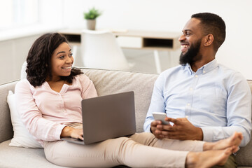 Fototapeta premium African american couple sitting on couch, using laptop and smartphone