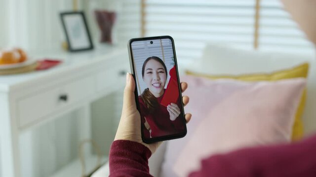 Point of view selfie videocall shot of attractive beautiful young asian lady in red chinese new year celebration outfit happy smile talk and looking to camera mobile phone wishing for lunar festival.