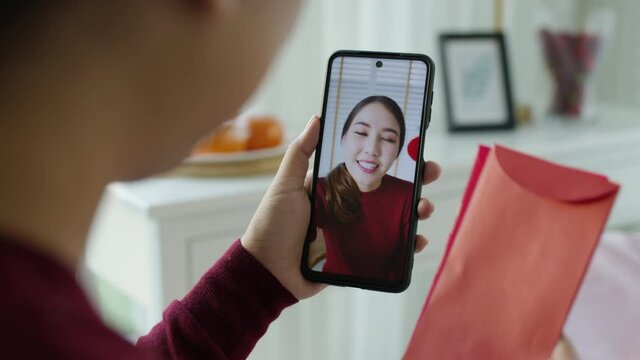 Point of view selfie videocall shot of attractive beautiful young asian lady in red chinese new year celebration outfit happy smile talk and looking to camera mobile phone wishing for lunar festival.
